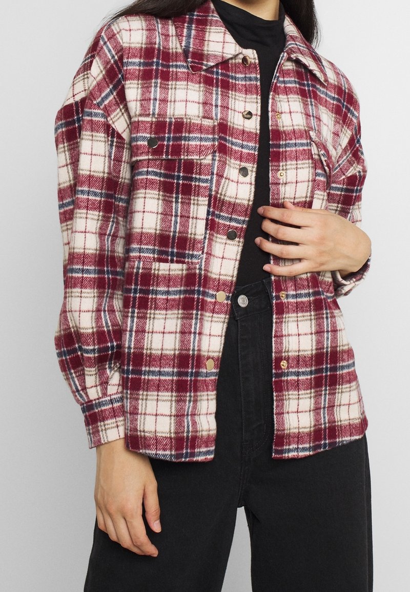 Personne portant une chemise à carreaux rouge, blanche et bleu marine boutonnée par-dessus un haut noir et un pantalon noir, la main reposant sur le ventre.