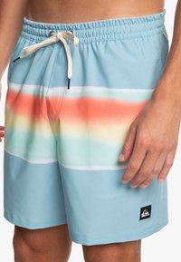 Pánske plážové šortky svetlomodrej farby s gradientným pruhovaným vzorom v oranžovej, žltej a zelenej. Majú elastický pás a šnúrku.