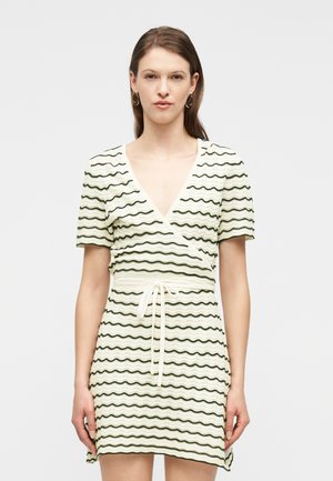 Mujer con cabello castaño liso que lleva un vestido cruzado de rayas onduladas beige y negro, con mangas cortas y cinturón de lazo, posando frente a un fondo blanco.