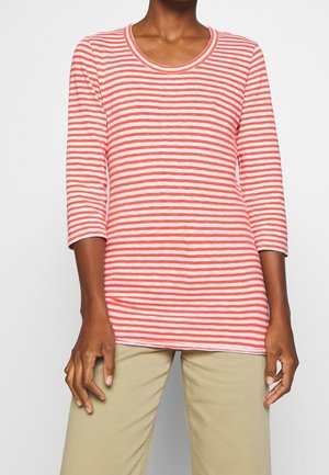 Person in rot-weiß gestreiftem Langarmshirt und beigen Hosen, zentriert vor einem schlichten hellgrauen Hintergrund.