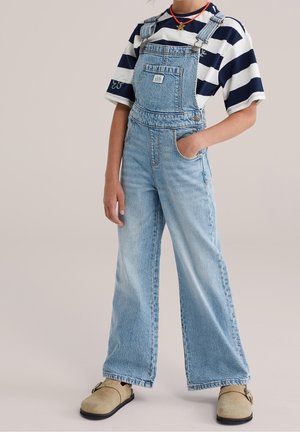Person iført lyseblå denim overalls, navy og hvide stribede kortærmede skjorte, tan slip-on sko og en stjerne halskæde.