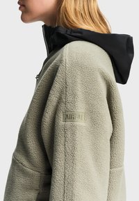 Woman wearing beige fleece jacket with black hood, visible AIGLE logo embroidered on upper arm, facing left in close-up side view.