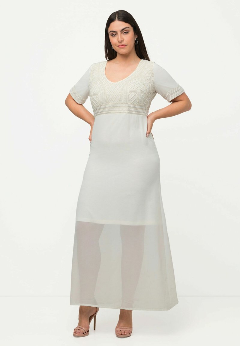 Maxikleid in hellem Creme mit kurzen Ärmeln und einem strukturierten bestickten Oberteil. Transparente Einsätze am Saum, ergänzt durch hellbeige Absätze.