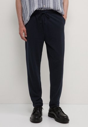 Mann trägt lockere marineblaue Hose mit Kordelzug, schwarze Schnürschuhe und ein gestreiftes Hemd mit Knöpfen, die Hände in den Taschen, steht vor einer weißen Wand.