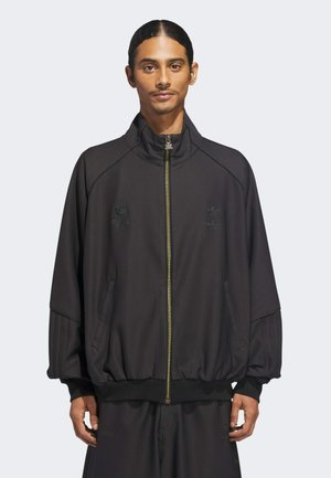Hombre con el cabello recogido, vestido con una chaqueta deportiva negra con cremallera y mangas acanaladas, y pantalones negros a juego, sobre un fondo claro y liso.