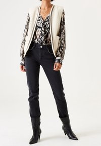 Krämfärgad faux fur-väst över en svartvit mönstrad blus, parat med svarta jeans och spetsiga svarta stövlar. Justerade ärmar med ränder.