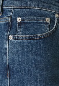 Jeans de mezclilla de color azul oscuro, con herrajes plateados y un pequeño bolsillo frontal. La textura es suave con un ligero desgaste.