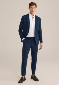 Homme portant un costume bleu marine ajusté avec une chemise blanche et des mocassins marron foncé, se tenant avec une main dans sa poche devant un fond uni.