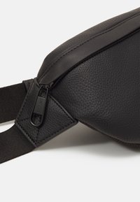 Musta nahkainen olkalaukku, jossa on teksturoitu pinta, sileä vetoketjusuljin ja säädettävä kangashihna turvallista käyttöä varten.