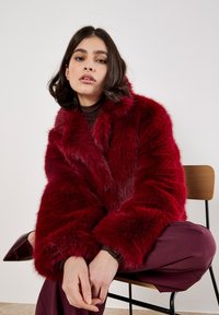 Junge Frau mit dunklen Haaren, die im Schneidersitz auf einem Stuhl sitzt und einen roten Kunstpelzmantel, bordeauxfarbene Hosen und spitze bordeauxfarbene Schuhe trägt.