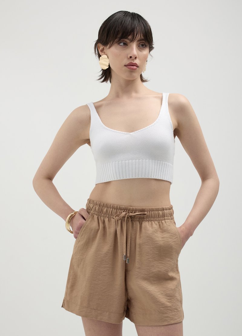 Haut court blanc à côtes avec un design en V, associé à un short beige à cordon de serrage. Le modèle se tient les mains dans les poches, mettant en valeur les détails de la tenue.