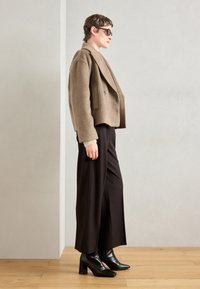 Persoon in een oversized beige wollen jas, zwarte wijde broek, zwarte haklaarzen en donkere zonnebril die zijwaarts staat op een houten vloer.
