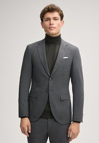 Grau karierter Blazer mit zwei Knöpfen, Reverskragen und seitlichen Taschen. Getragen über einem dunklen Rollkragenpulli, mit einem weißen Einstecktuch.