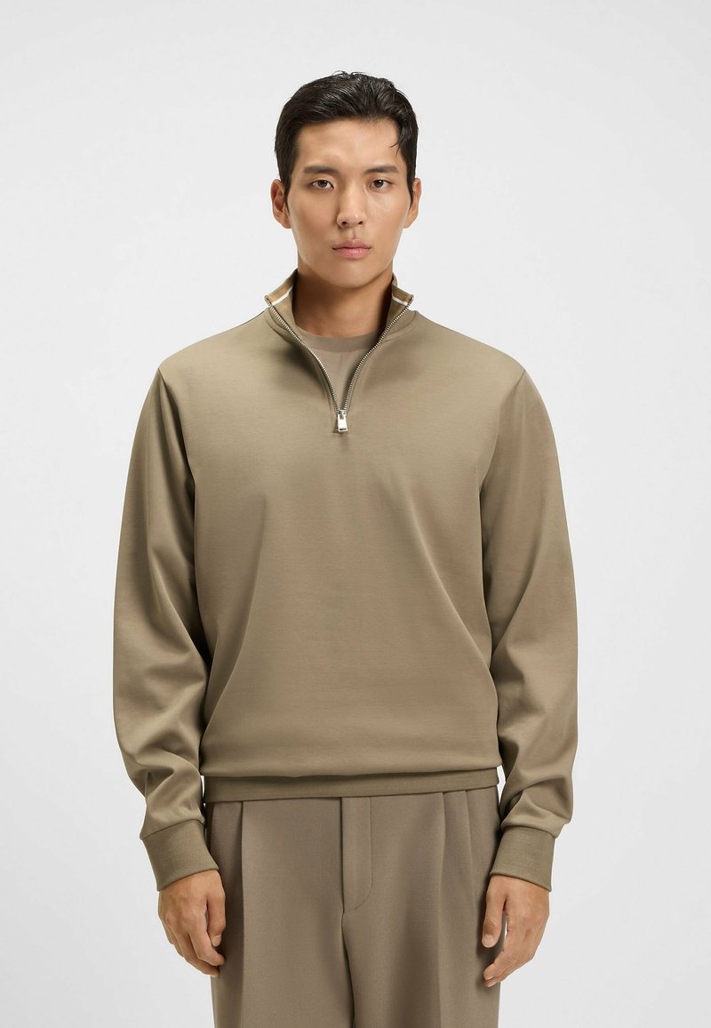 Beige zip-up pullover made of soft fabric with a stand collar. Features long sleeves and a relaxed fit. Textured material with subtle sheen.