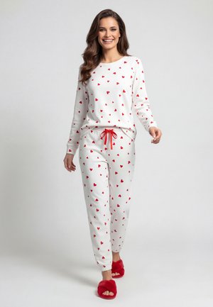 Vrouw die loopt in een wit pyjamaset met rode hartjes, rode trekkoorden en rode pluizige pantoffels, glimlachend tegen een effen lichte achtergrond.