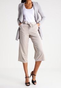 Culottes beiges taille haute avec des détails de ceinture nouée, fabriqués en tissu lisse. Associés à un débardeur blanc et à un cardigan texturé gris.