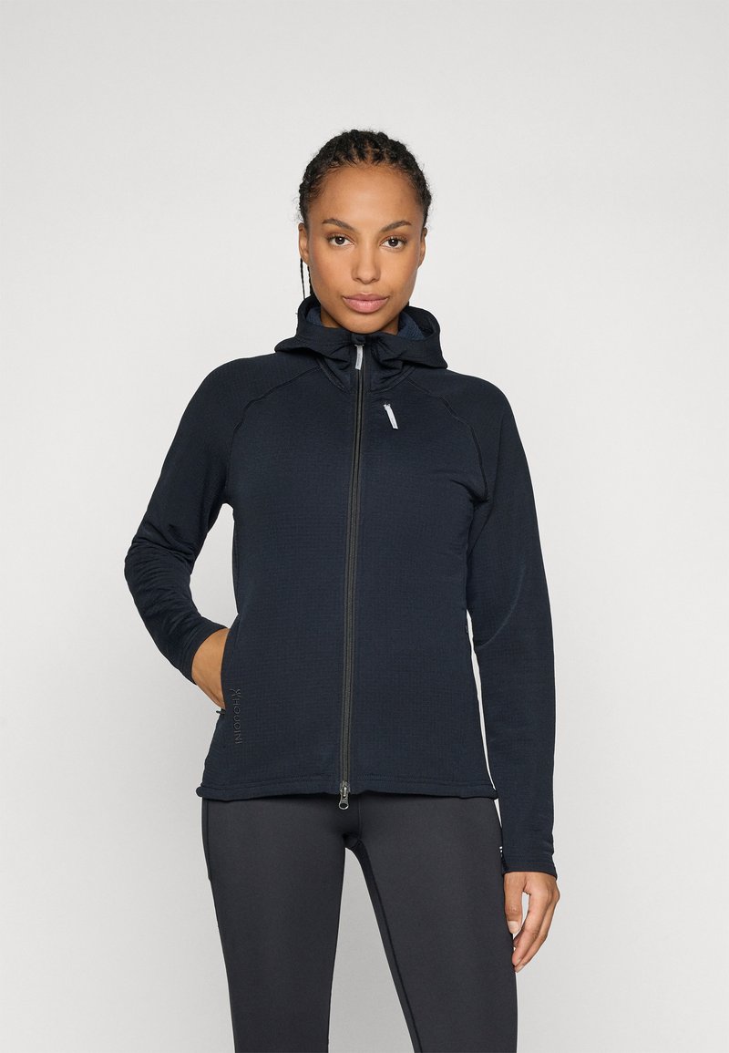 Dunkelblaues Zip-Up-Hoodie mit tailliertem Schnitt, strukturiertem Stoff und Kapuze. Mit einem Frontreißverschluss und seitlichen Taschen. Kombiniert mit schwarzen Hosen.