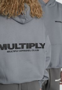 Sivá mikina s kapucňou s veľkým čiernym logom "MULTIPLY" na chrbte a textom "MULTIPLY APPAREL CLUB". Mäkký materiál, voľný strih.