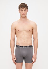 Sive bokserice s crnim pojasom na kojem je logo "BJÖRN BORG". Glatka tekstura i oblikovani dizajn. Model stoji s neutralnim izrazom lica.