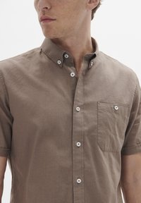 Cămașă maro cu mânecă scurtă, fabricată dintr-un material țesut; are un guler tip button-down, un buzunar frontal și nasturi albi. Textura pare netedă.