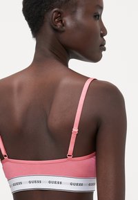 Ružičasti bralet s tankim podesivim trakama, koji ima bijeli pojas s "GUESS" logotipom. Glatka tekstura i prilagođen dizajn ističu leđa.