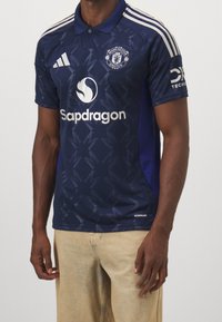 Granatowa koszulka polo z krótkimi rękawami, z wyrazistym wzorem geometrycznym, logo Adidas oraz napisem "Sapdragon" z przodu.