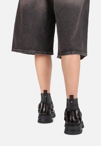 Bottines en cuir verni noir avec des côtés texturés, des boucles et des semelles épaisses. Portées avec un short en denim gris.