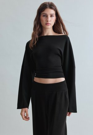 Ung kvinde med langt brunt hår iført en sort off-shoulder langærmet top og sorte bukser mod en ensfarvet lys baggrund.