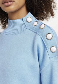 Jersey de color azul claro con un alto cuello acanalado, que presenta botones decorativos blancos y detalles metálicos en el hombro. Tejido de textura suave.