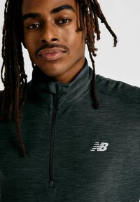 Veste zippée noire avec texture et col haut ; présente un logo New Balance blanc sur la poitrine ; vue en gros plan du haut du corps d'une personne.