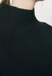 Gebreide coltrui in donker groen, met een geribbelde textuur en hoge kraag, met diagonale lijnen in de stof.