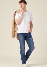 Weißes kurzärmliges T-Shirt mit einer Brusttasche, kombiniert mit blauen Jeans und einer beigefarbenen Jacke, die über die Schulter geworfen ist. Weiße Sneaker.