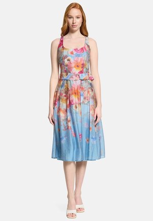 Kobieta w sukience midi bez rękawów, z paskiem, ozdobionej dużym różowym, pomarańczowym i niebieskim kwiatowym wzorem na jasnoniebieskim tle, w białych sandałach.
