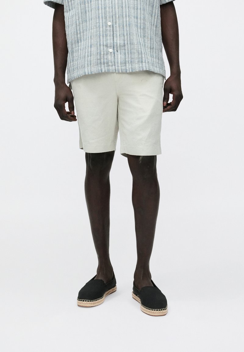 Lichtgekleurde linnen shorts hebben een losse pasvorm en worden gecombineerd met een contrasterend, gestructureerd grijs-blauw gestreept overhemd en zwarte espadrilles met gevlochten zolen.