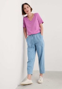 Růžové tričko s výstřihem do V, světlomodré kalhoty s elastickým pasem a kapsami a krémové tenisky, na pozadí jednoduché zdi.