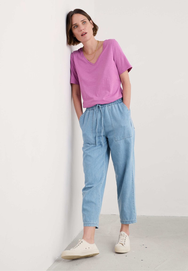 Růžové tričko s výstřihem do V, světlomodré kalhoty s elastickým pasem a kapsami a krémové tenisky, na pozadí jednoduché zdi.