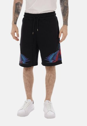Uomo che indossa pantaloncini neri con disegni di ali di piume blu e rosse sulle cosce, scarpe da ginnastica bianche, camicia bianca e tatuaggi visibili sulle braccia.