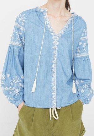Femme portant un chemisier bleu clair brodé avec des liens à pompons et des manches bouffantes, associé à un pantalon vert olive.