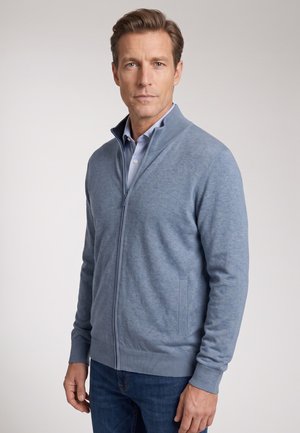 Mann trägt einen hellblauen Zip-up-Pullover über einem Hemd mit Kragen und dunkelblaue Jeans, steht vor einem einfarbigen hellen Hintergrund.