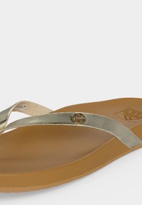 Καφέ σαγιονάρες με μεταλλικές ασημένιες λουρίδες και ένα μικρό στρογγυλό χρυσό έμβλημα στο πλαϊνό λουράκι, σε μια απλή λευκή επιφάνεια.