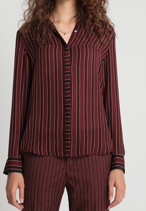 Woman wearing a burgundy long-sleeve blouse and pants with vertical black and pink stripes, standing against a plain background.