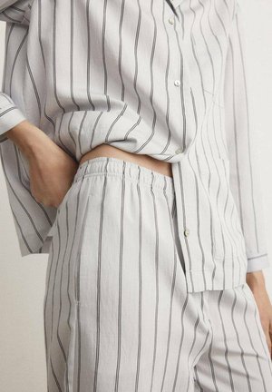 Person wearing white pajamas with thin black vertical stripes, one hand tucked into the elastic waistband of the pants.