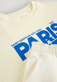 Creme sweatshirt med blå "PARIS" grafik, ribbet halsudskæring. Blødt, let stof. Creme og blegt stribet stofdetalje i forgrunden.