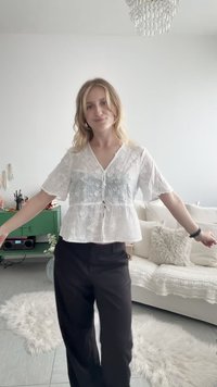Femme aux cheveux blonds portant un haut floral blanc transparent et un pantalon noir, debout les bras légèrement écartés dans un salon décoré.