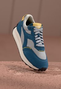 Blue and gray sneaker with suede and mesh panels, rounded toe, white sole, gray laces, and yellow accents on the tongue and stitching.