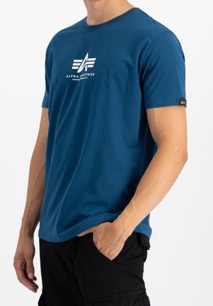 Mężczyzna w niebieskiej koszulce Alpha Industries z białym logo i czarnych spodniach typu cargo, z jedną ręką w kieszeni, na jednolitym białym tle.