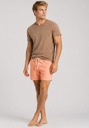Mand stående barfodet iført en brun kortærmet skjorte og koralfarvede shorts med snøre, kigger til siden mod en ensfarvet baggrund.