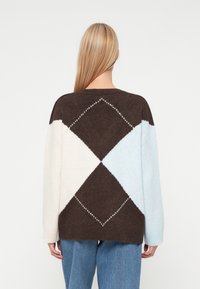 Γυναίκα με μακριά ξανθά μαλλιά, φορώντας ένα καφέ, λευκό και ανοιχτό μπλε πουλόβερ με σχέδιο αργάκη και μπλε τζιν, στραμμένη μακριά από την κάμερα.