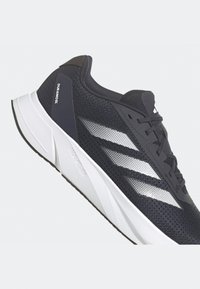 Chaussure de running Adidas noire et blanche avec une semelle intermédiaire Duramosi et trois bandes blanches sur le côté, inclinée pour montrer le côté extérieur et les lacets.