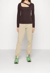 Brązowy prążkowany longsleeve z prostokątnym wycięciem, w zestawie z beżowymi spodniami dresowymi i zielonymi butami sportowymi. Widoczne detale strukturalne materiału.